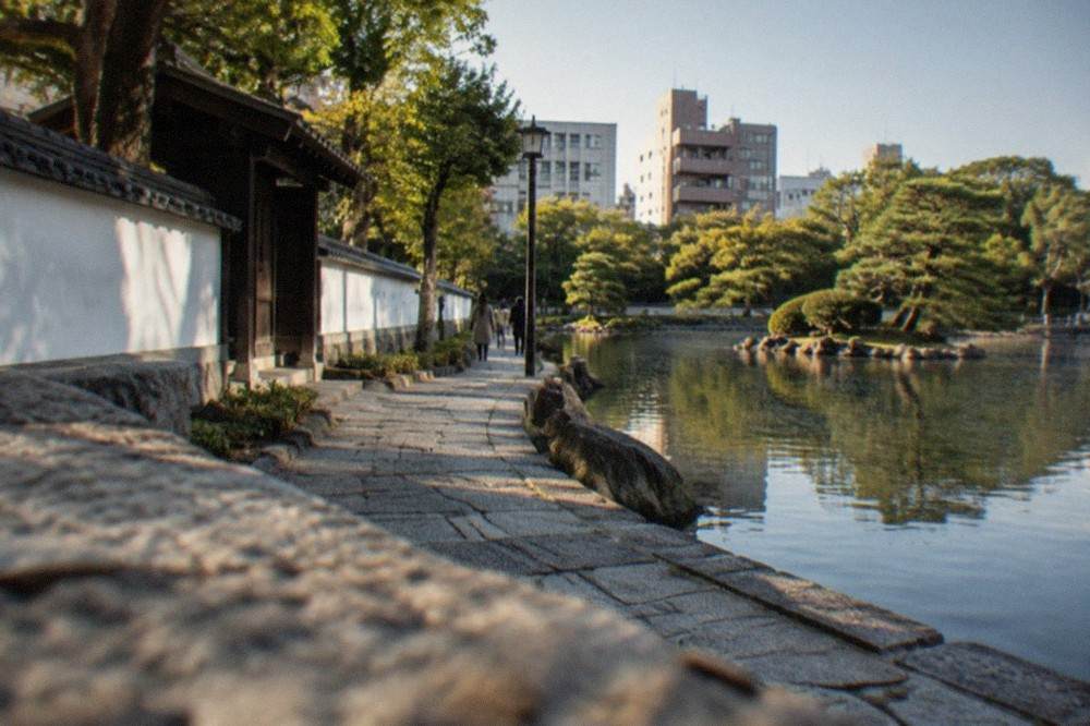 A stone walking path beside a large pond with a traditional garden in the background