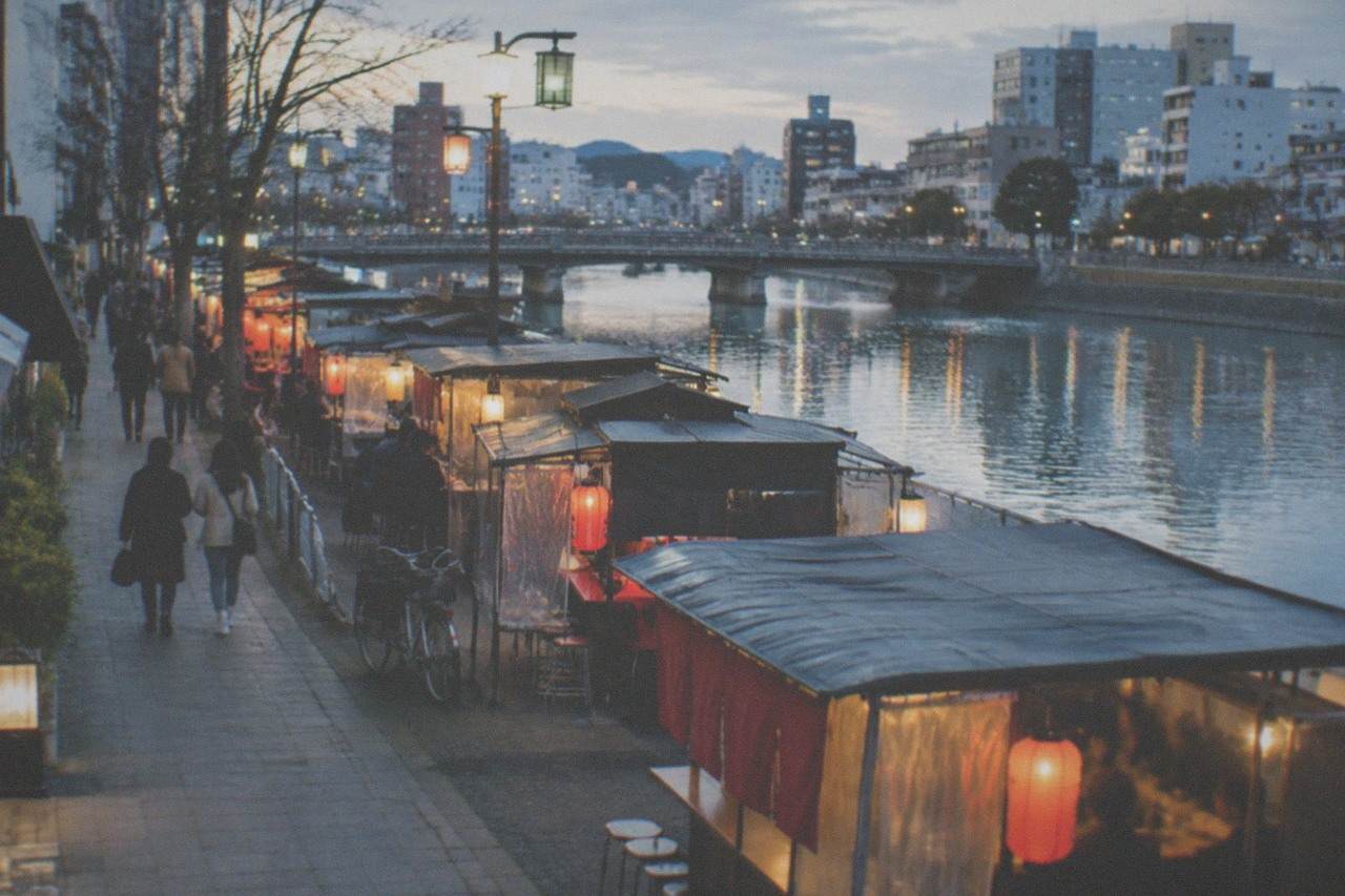 Evening food stalls with lanterns glowing along a riverside street in a Japanese city