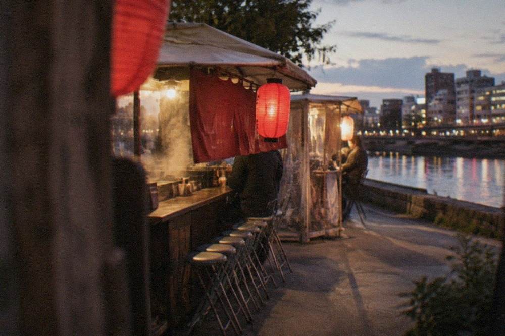 A lit food stall with red lanterns glowing beside a canal at night in a Japanese city