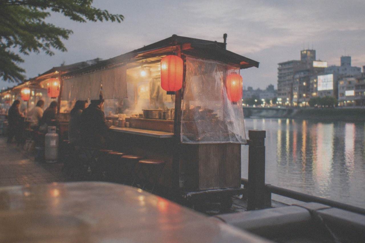 A lit food stall with red lanterns glowing beside a canal at night in a Japanese city