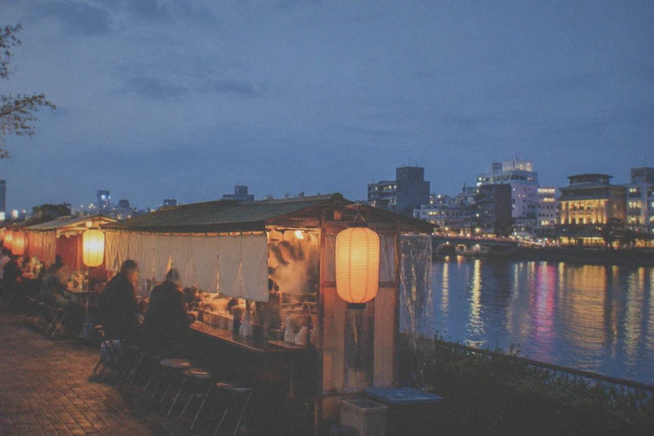 A row of glowing yatai food stalls along the Naka River at dusk, with city lights reflecting on the water.