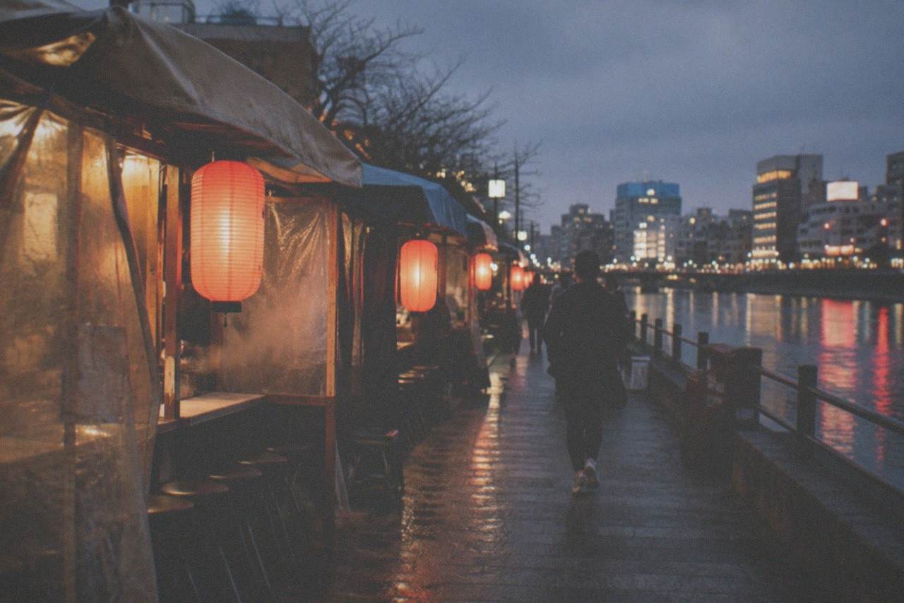 A row of glowing food stalls lined up along a river at night, with red lanterns reflecting on the water