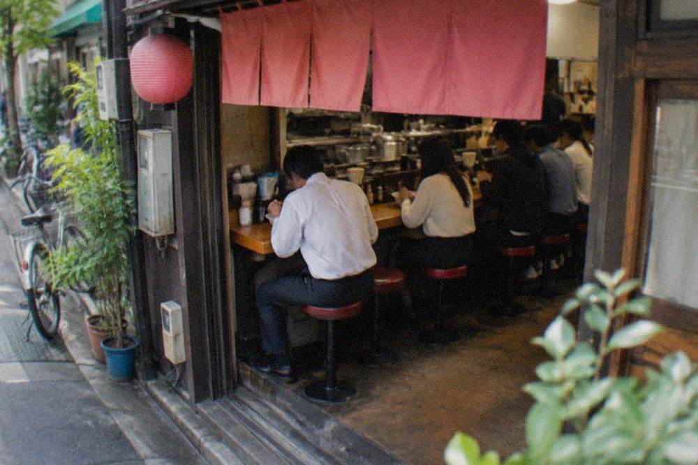 A narrow ramen shop counter with diners seated side by side eating bowls of noodles