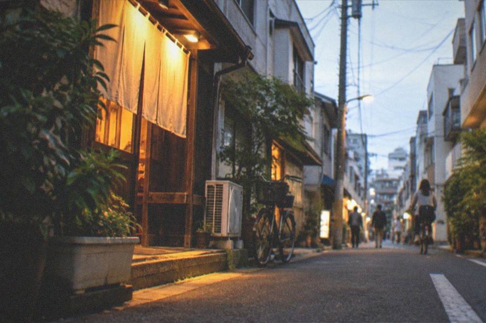 An evening street corner with warm light spilling from a small shop entrance onto the pavement