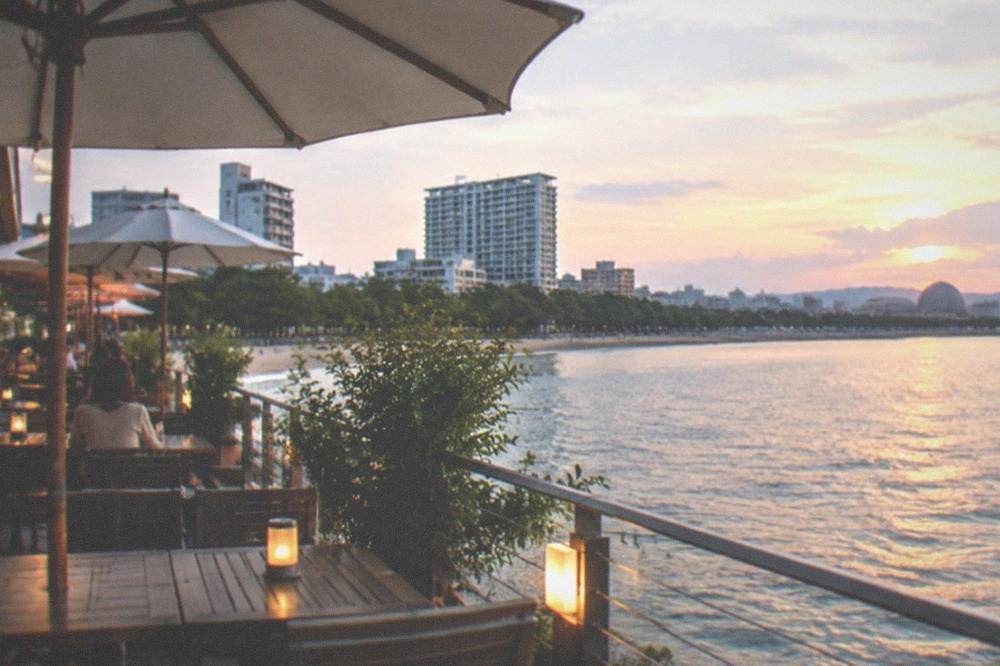 A waterfront restaurant terrace with umbrella tables overlooking a calm bay at dusk
