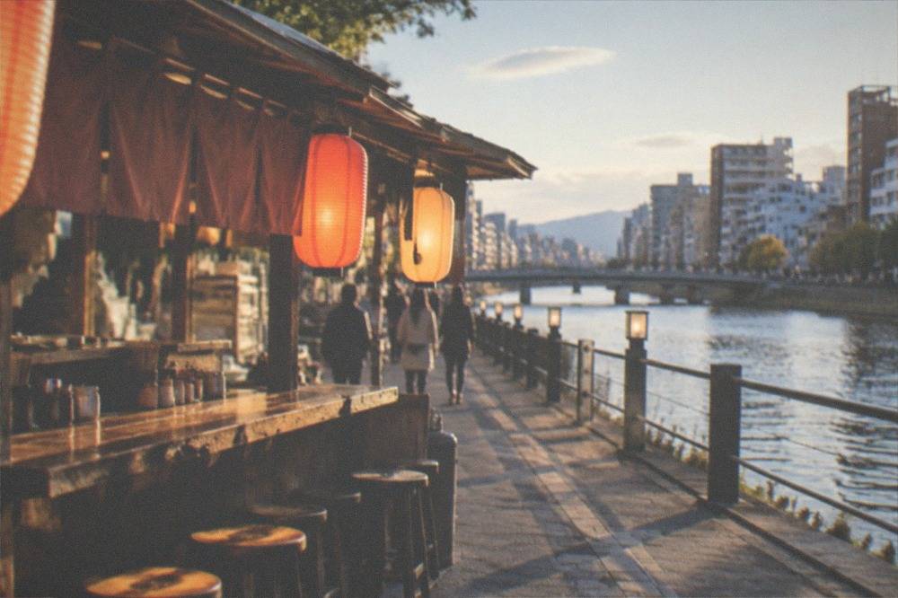 A small wooden food stall with glowing lanterns and a counter of stools beside a river