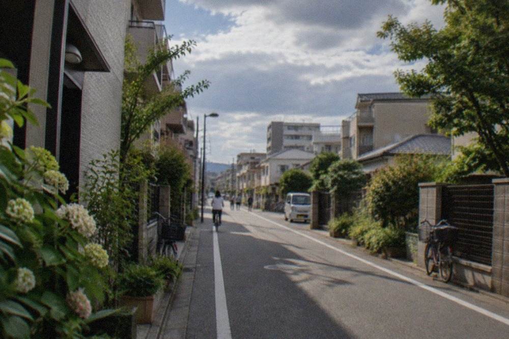 A quiet residential street in Nishijin with low-rise buildings and greenery along the sidewalk