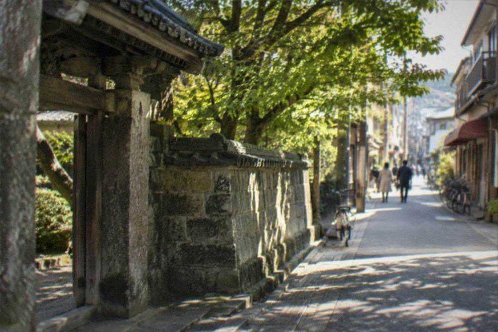 A weathered stone gate and wall of an old temple beside a narrow tree-shaded street