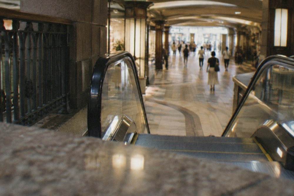 Escalators descending into a brightly lit underground concourse with tiled floor and shopfronts