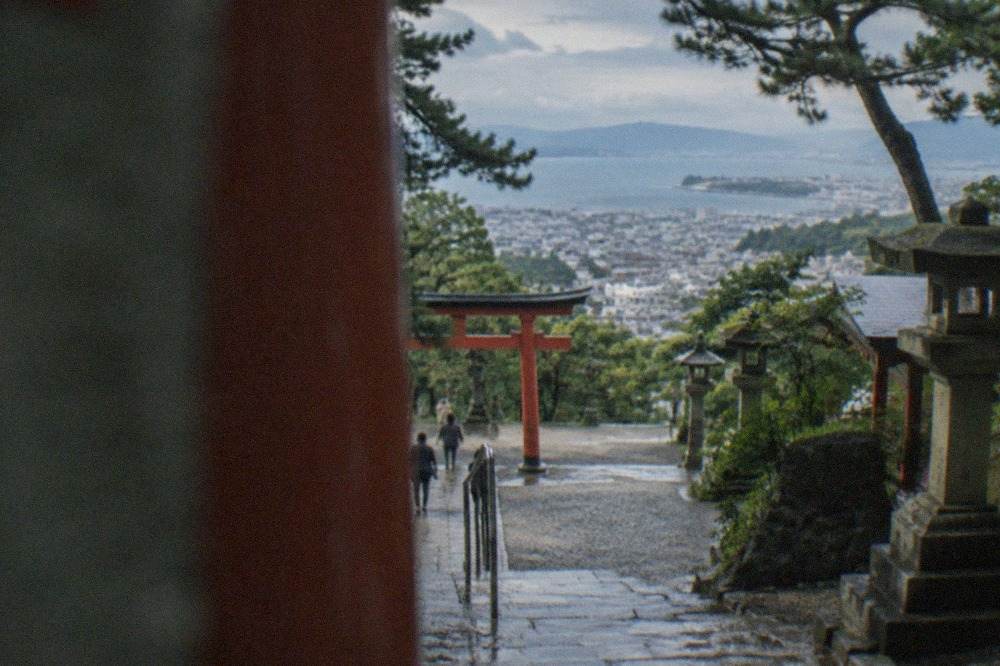 Stone steps rising through trees toward a vermilion shrine gate on a wooded hillside above the city