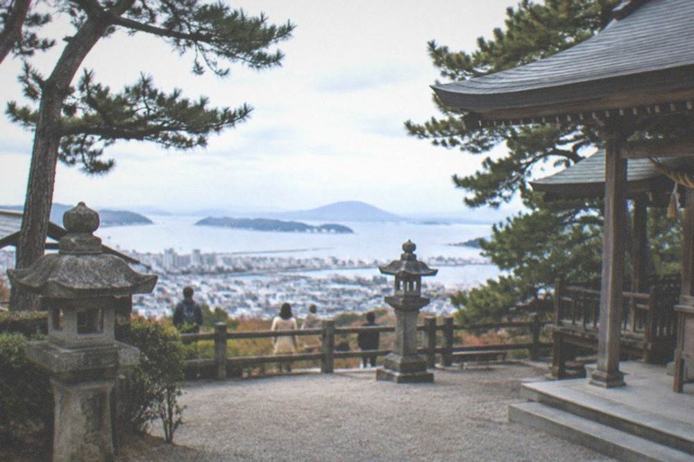 A hilltop clearing with a wooden shrine hall and a view of a bay and distant islands beyond the trees