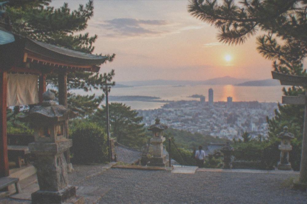 A city skyline of glass towers beside a wide bay at sunset with islands on the horizon