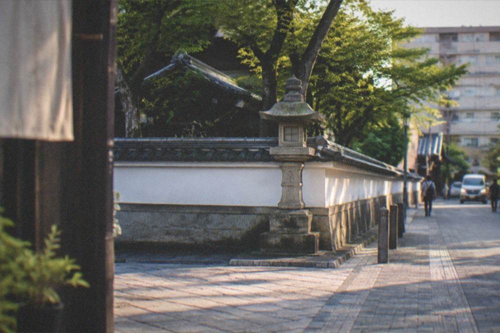 A quiet stone-paved street corner with a temple wall and lantern in soft evening light.
