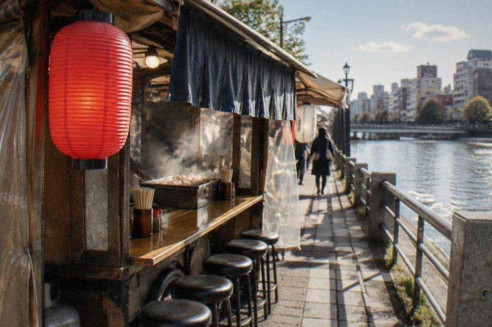 Close view of a small food stall cart with a red lantern and a narrow lit counter beside a river path