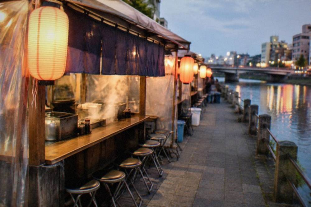 Bridge over a city river at night with warm stall lanterns glowing along the far bank