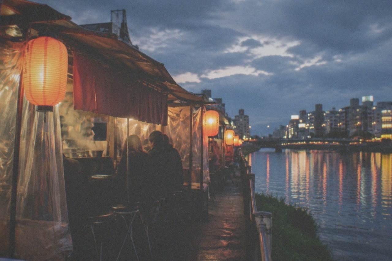 Row of glowing food stalls lined up along a river at night, with lanterns reflected in the water
