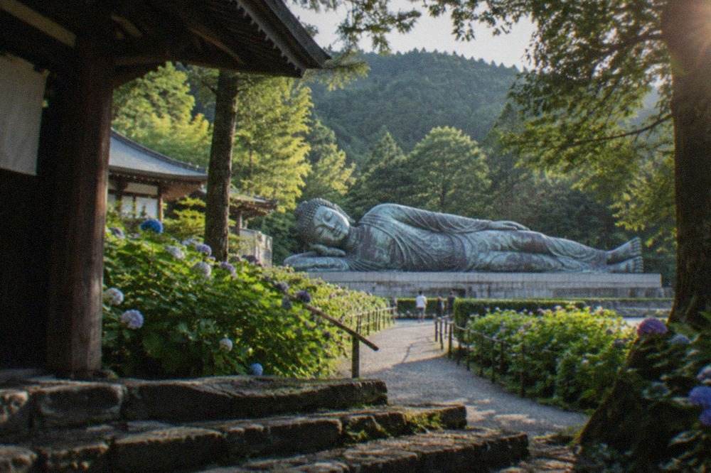 A very long bronze reclining Buddha statue lying in an open plaza surrounded by pine-covered hills