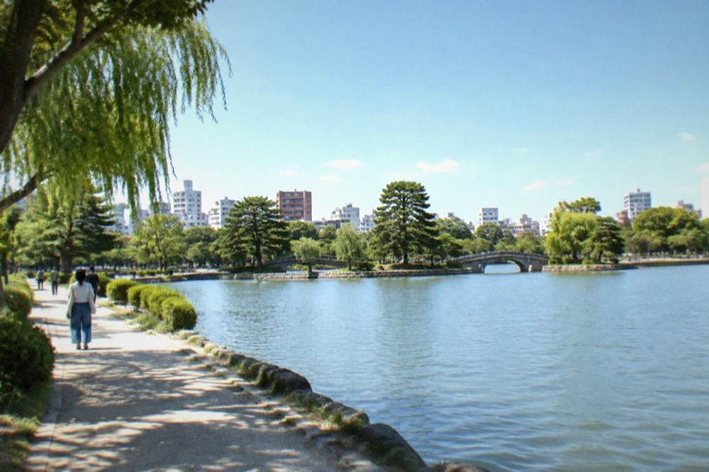 A tree lined walking path beside pond water with city buildings beyond