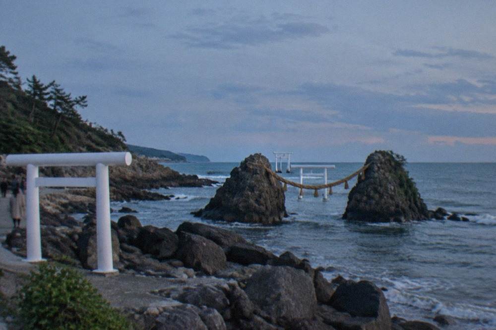 Two sea stacks joined by a thick rope, with a white torii gate standing in the water behind them under a wide evening sky