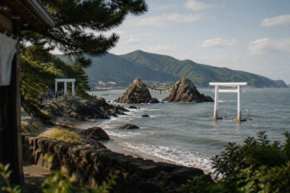 A white torii gate standing in shallow sea water with the coast and green hills of Itoshima behind it