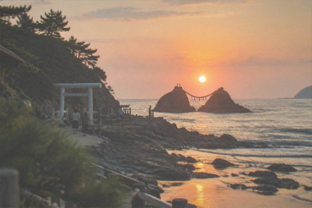 The sun setting behind two sea stacks and a torii gate, with orange light across the sea and wet sand reflecting the sky