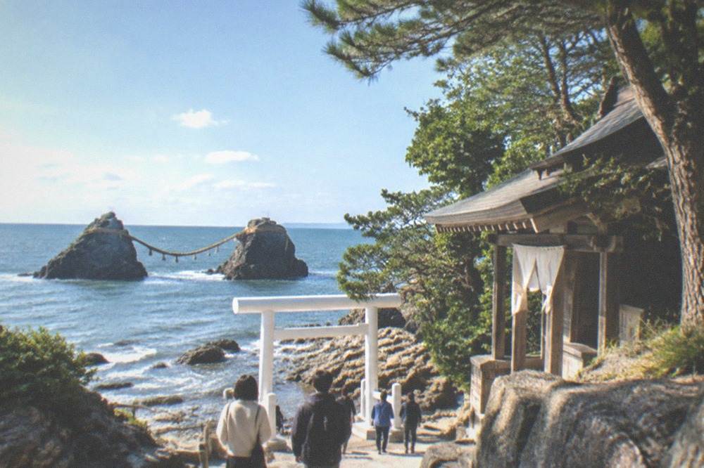 A small shrine building with wooden details standing among trees near the coast