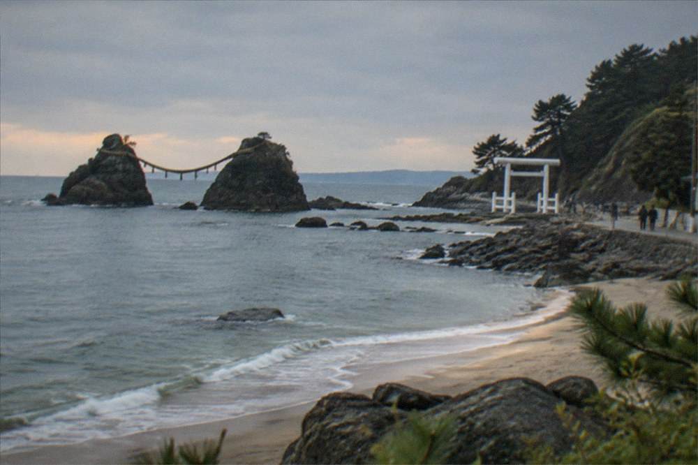 A calm beach at dusk with soft light on the sand and dark rocks visible out in the sea