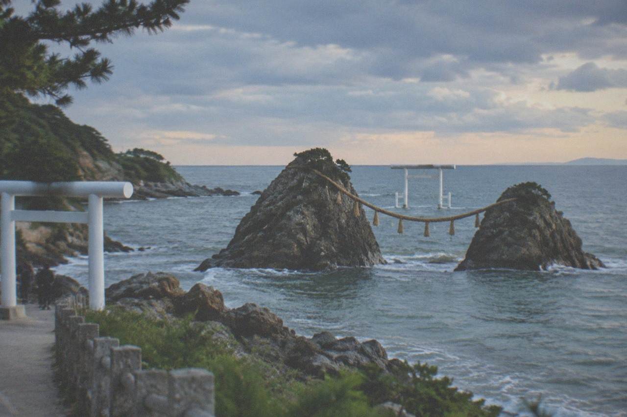Two sea stacks joined by a thick rope, with a white torii gate standing in the water behind them under a wide evening sky