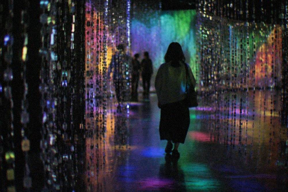 A visitor walking through a corridor of hanging mirrored strands lit in shifting colours