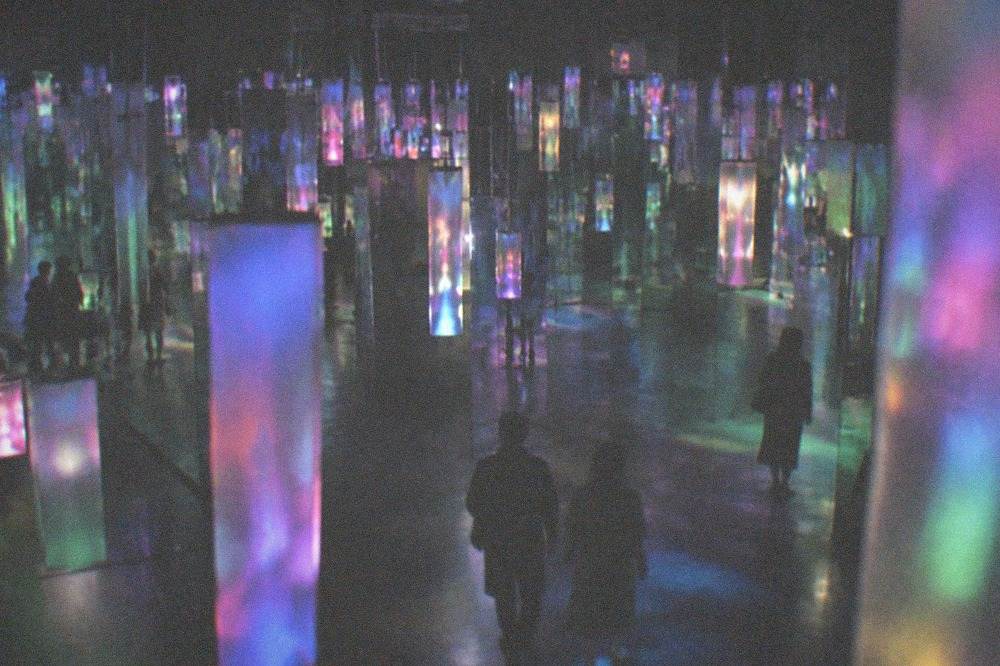A dark gallery space filled with hanging lights and mirrored surfaces reflecting colour