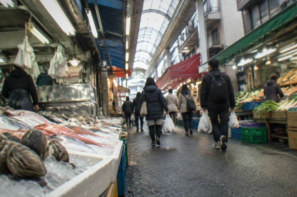 Narrow covered arcade aisle with shop awnings and shoppers carrying bags in Fukuoka