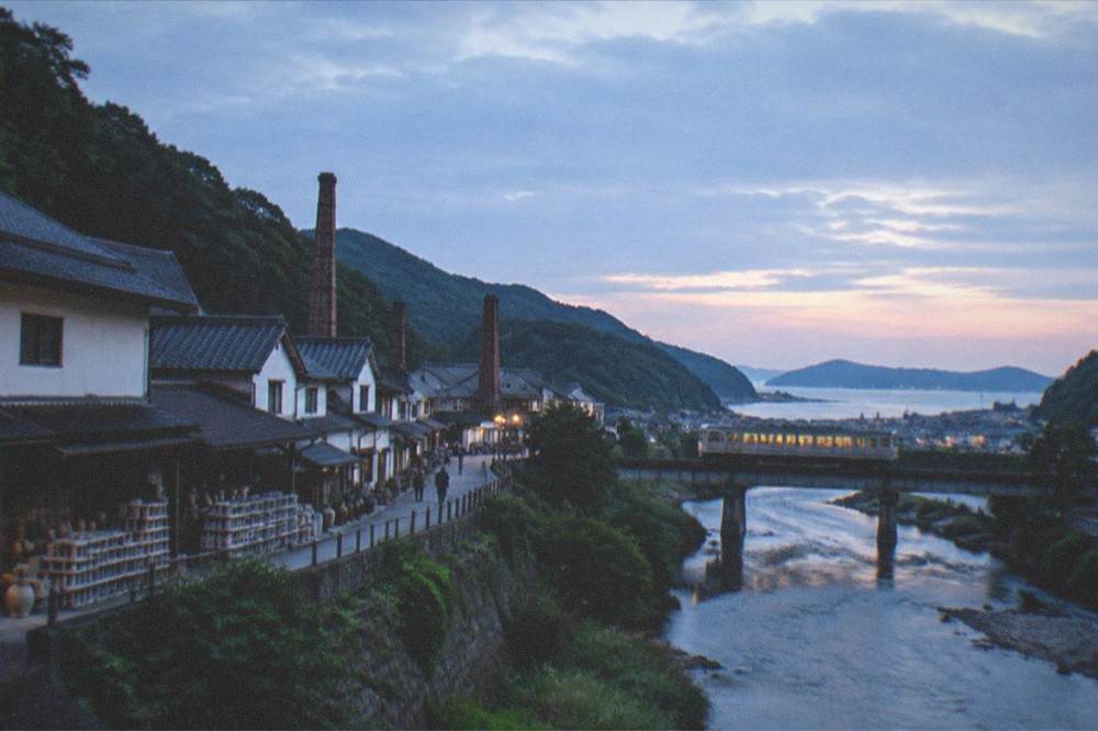 A train crossing a river bridge through green hills toward a coastal town in evening light