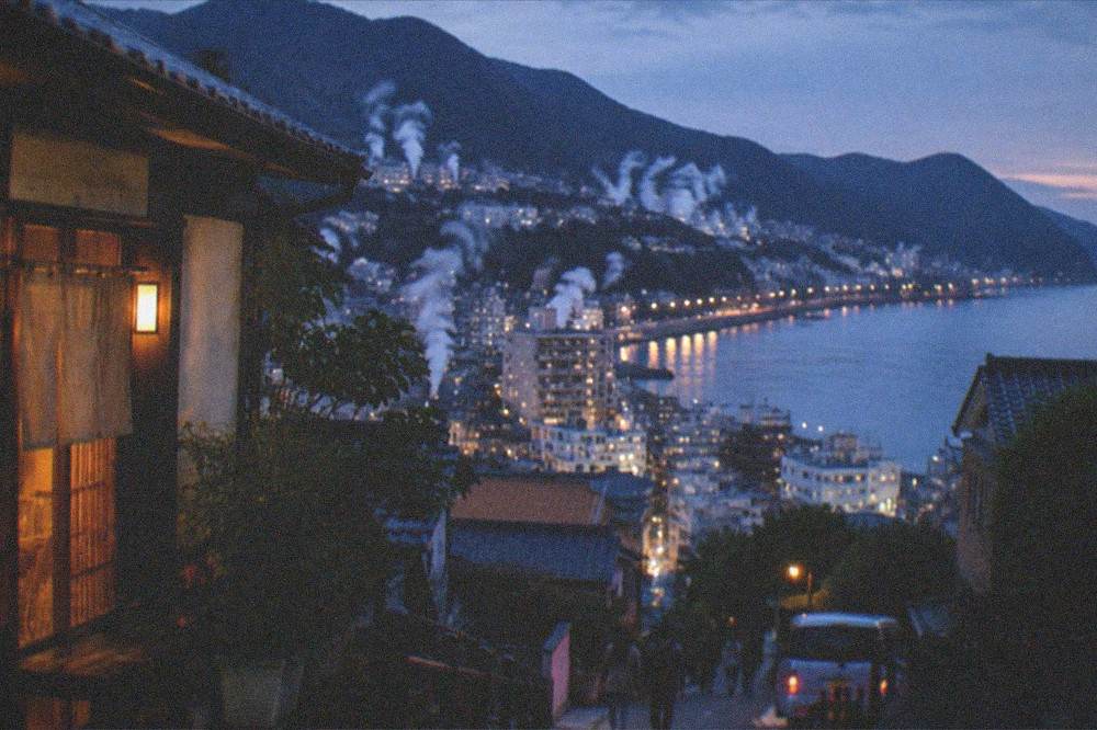 A coastal town at dusk with lights along the waterfront and hills behind