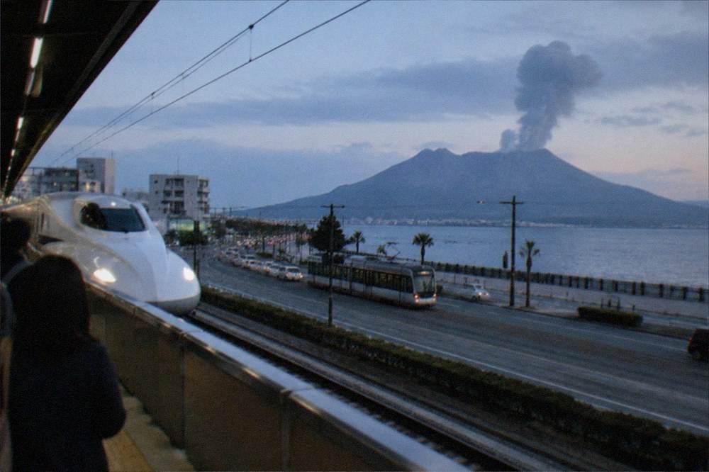 A station platform with a white bullet train nose visible and travellers waiting behind a yellow safety line