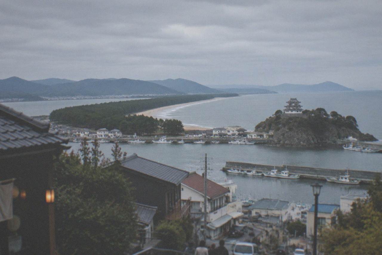 A Japanese castle keep on a hill above a wide bay, with pine-covered sandbar curving along the shoreline in the distance