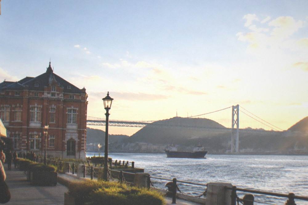 Red-brick harbour buildings with white stone trim facing a strait with a suspension bridge behind