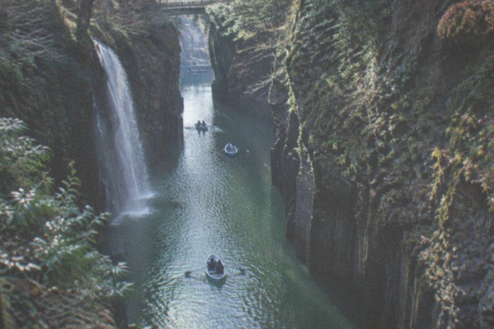 Small rowing boats moving slowly along a calm green river between tall basalt cliffs