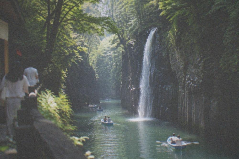 A waterfall pouring over a dark cliff edge into a narrow forested gorge in summer
