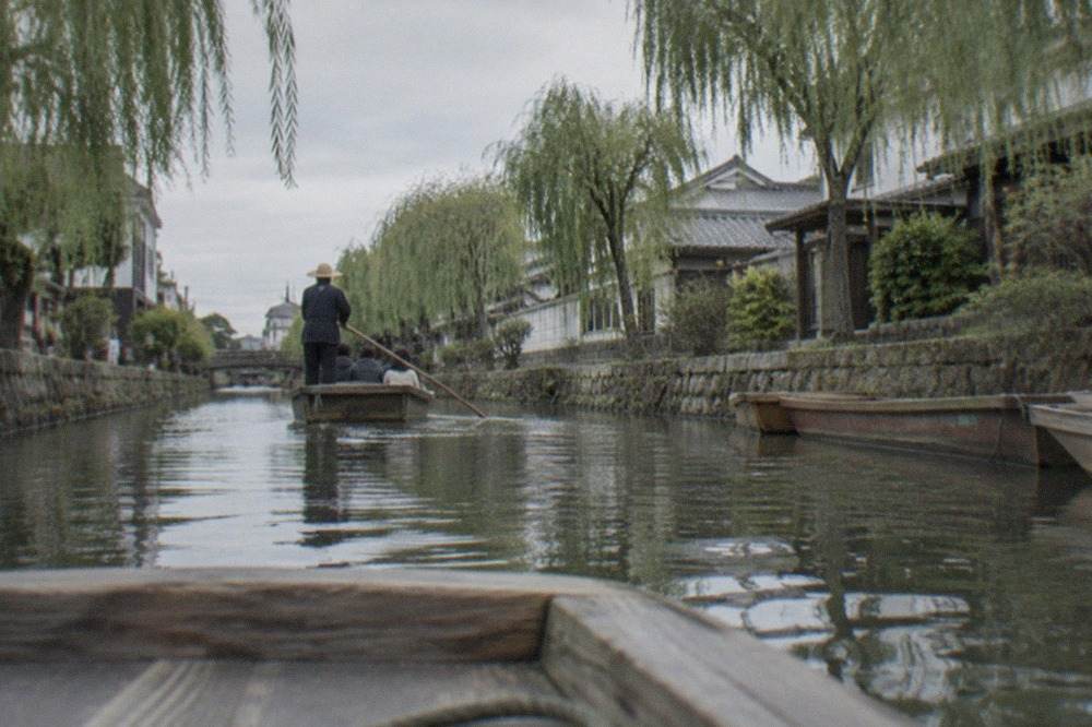A flat wooden punt boat gliding along a narrow canal lined with stone walls and willow trees