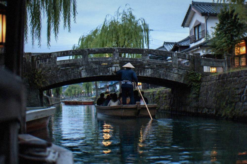 A boatman in traditional work clothes poling a punt under a low stone bridge while passengers duck