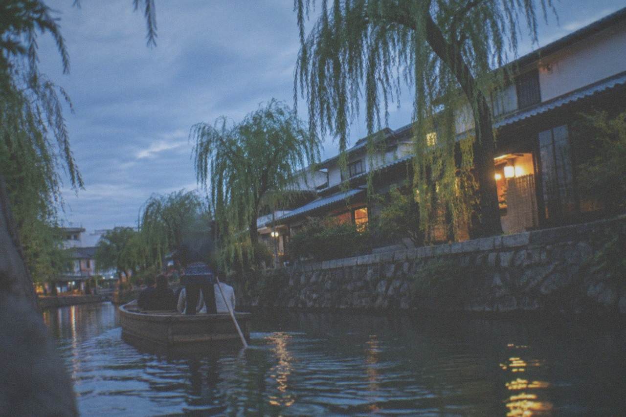A flat wooden punt boat gliding along a narrow canal lined with stone walls and willow trees