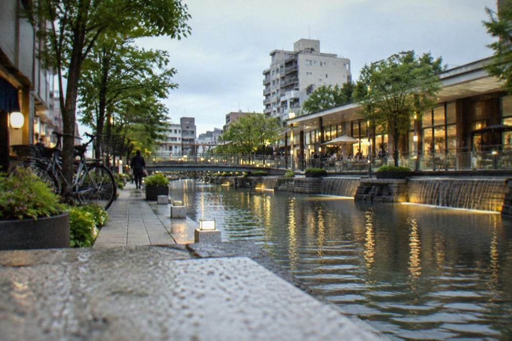 A modern canal-side shopping complex with illuminated walkways and water features in the evening