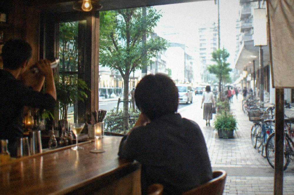 A bartender shaking a cocktail at a wooden counter with a single customer seated