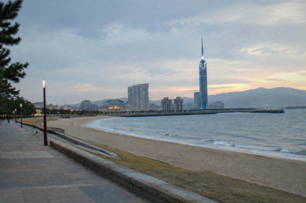 A seaside promenade at dusk with a tall illuminated tower glowing over a darkening bay