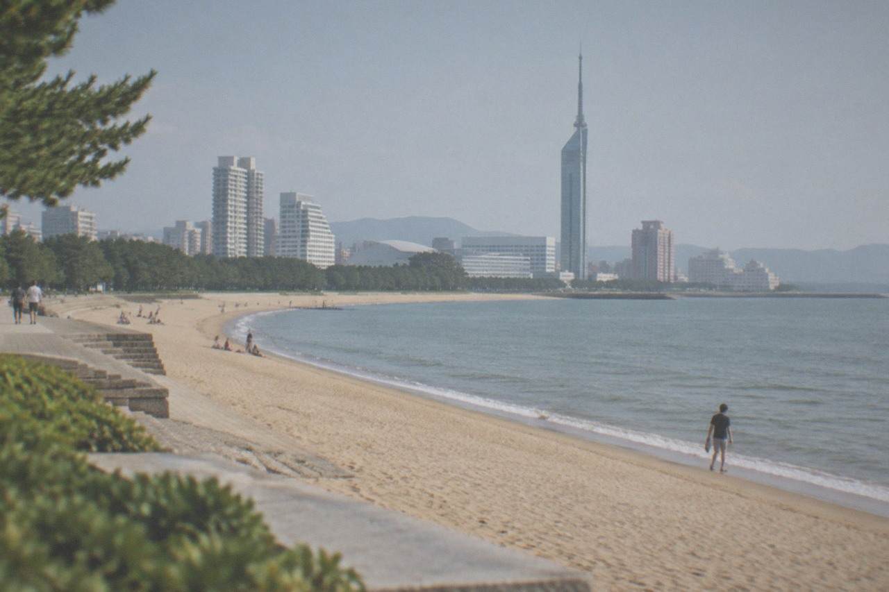 A wide sandy beach curving along a calm bay with a tall seaside tower in the background under a clear sky