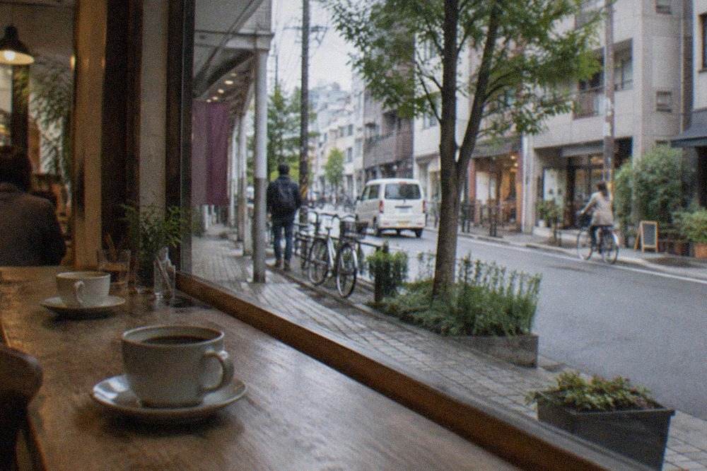 A quiet cafe interior with wooden tables, cups of coffee on a counter, and a window onto a city street
