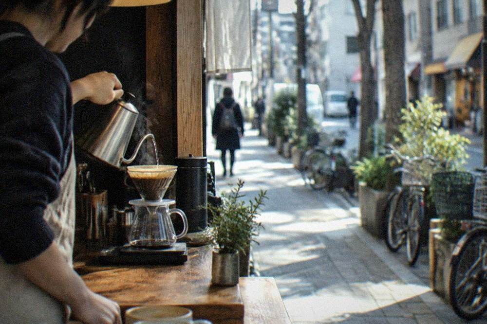 A barista pouring hot water over a coffee filter behind a small wooden counter