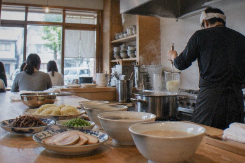 A small teaching kitchen with a wooden counter, bowls of pork broth, noodles and toppings set out ready for a class