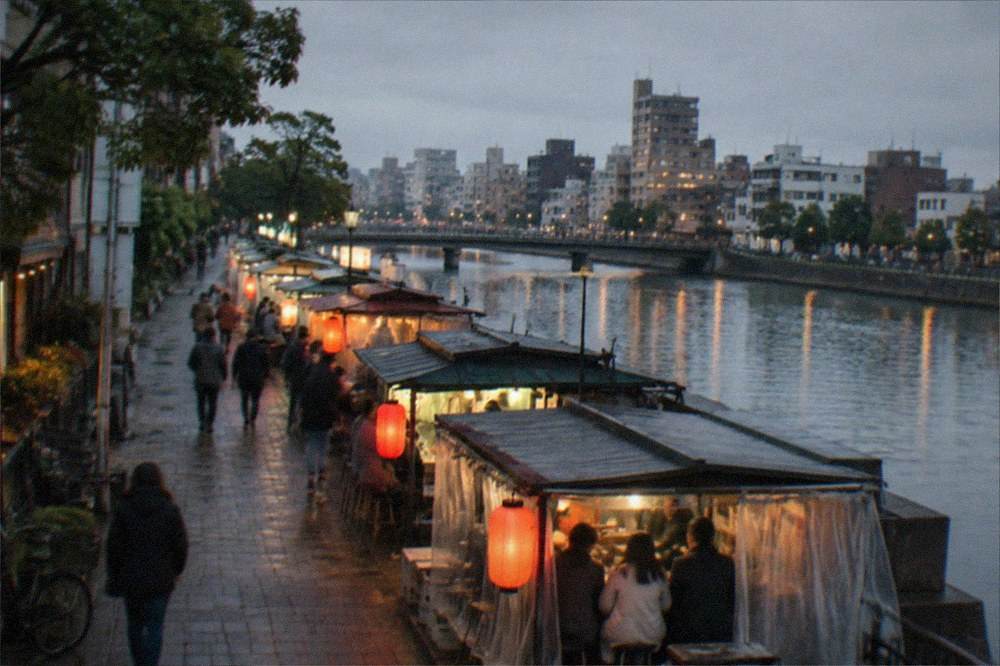 A row of illuminated street food stalls lining a riverside street after dark