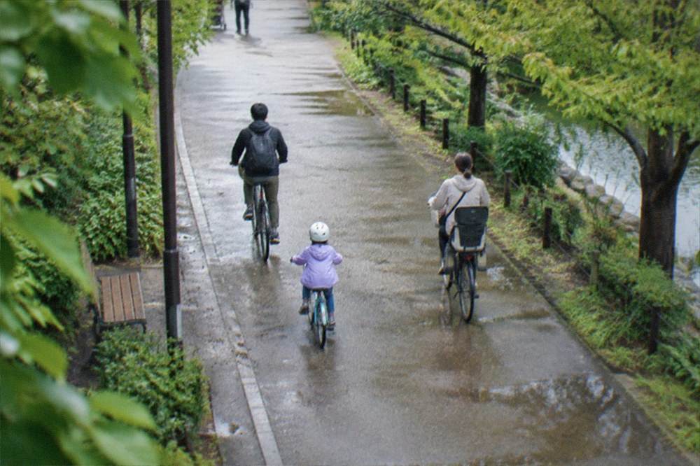 A family cycling together on a wide flat path through parkland in Fukuoka